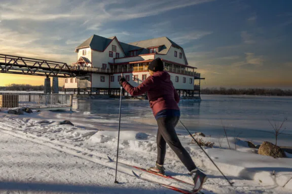 Au crépuscule, personne arrivant à la hauteur de la Maison riveraine en ski de fond, en longeant la rivière des Outaouais gelée.
