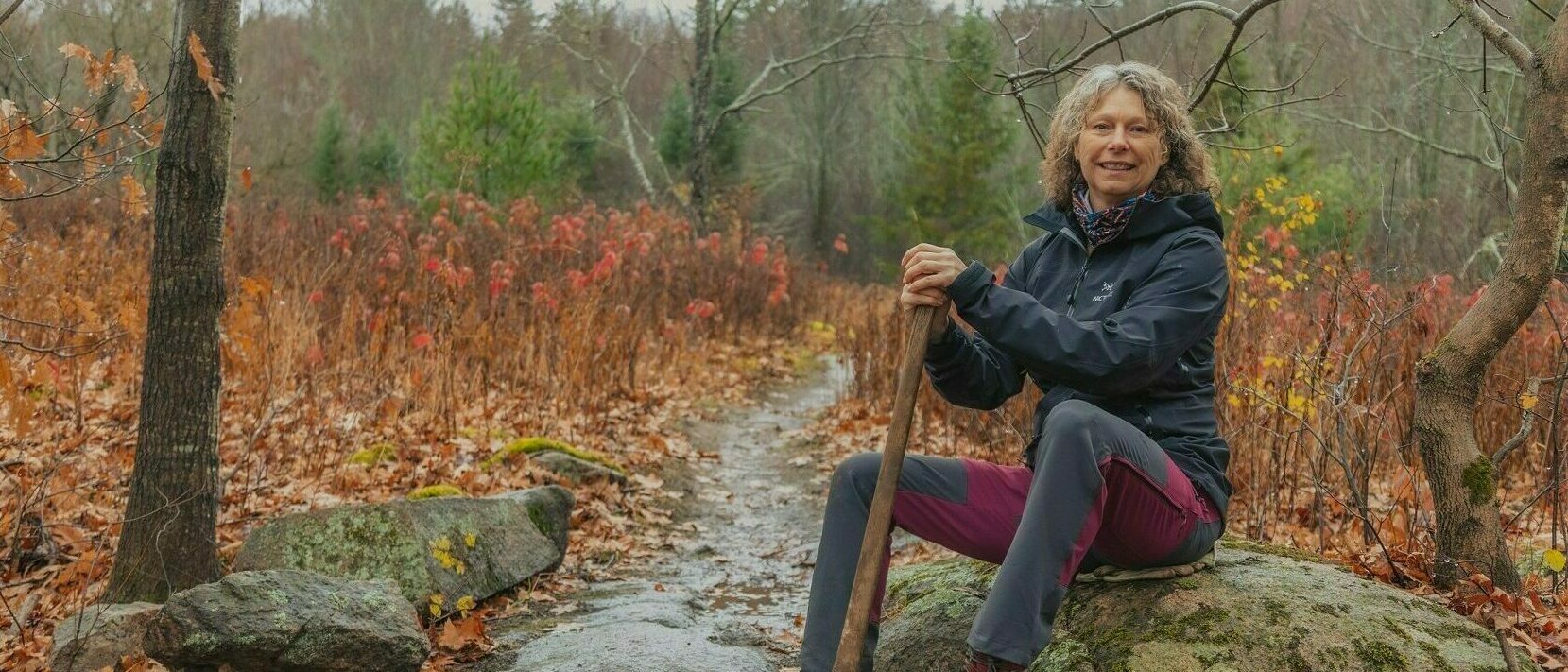 Volunteer sitting on a rock, facing the camera, at a trail entrance in Gatineau Park.